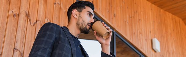 Vista de bajo ángulo del empresario árabe bebiendo café cerca del edificio con fachada de madera, pancarta  - Foto, imagen