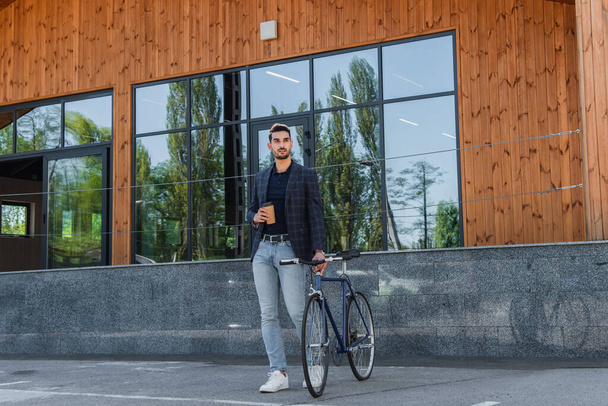 Jovem gerente árabe segurando copo de papel perto de bicicleta e construção  - Foto, Imagem