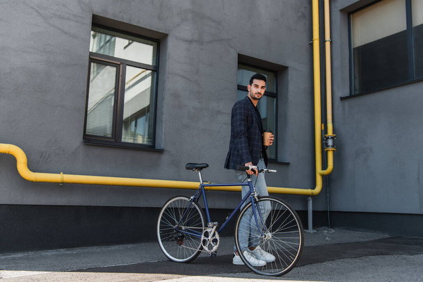 Arabian businessman with paper cup looking at camera near bike outdoors  - Fotoğraf, Görsel