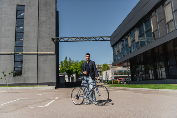 Arabo uomo d'affari con caffè e bicicletta sorridente alla macchina fotografica sulla strada urbana  - Foto, immagini
