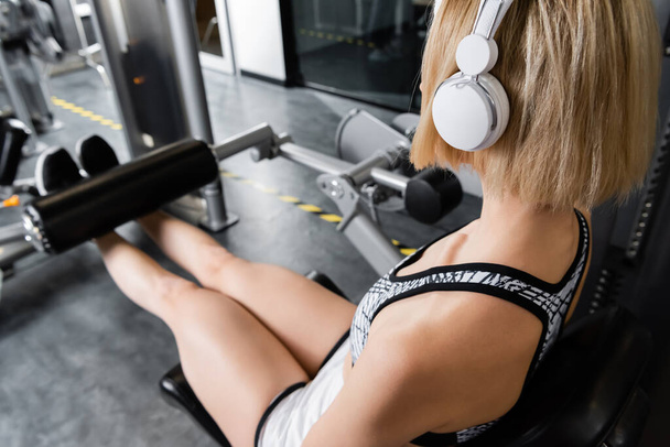 mujer joven en el entrenamiento de auriculares inalámbricos en la máquina de ejercicio de extensión de pierna - Foto, imagen