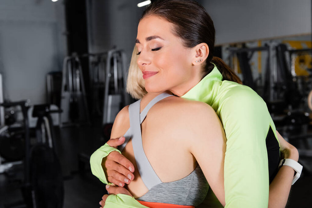 χαρούμενη γυμνάστρια με κλειστά μάτια που αγκαλιάζει αθλήτρια στο γυμναστήριο - Φωτογραφία, εικόνα