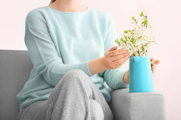 Женщина с кувшином красивых цветущих ветвей на диване в комнате - Фото, изображение