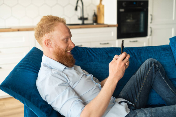 Serene smiling young redhead man lying down on the sofa with a smartphone and hands - Foto, Bild