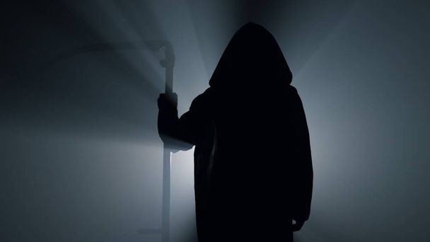 Η Silhouette Grim Reaper περιμένει με δρεπάνι μέσα. Scytheman στέκεται σκοτάδι. - Φωτογραφία, εικόνα