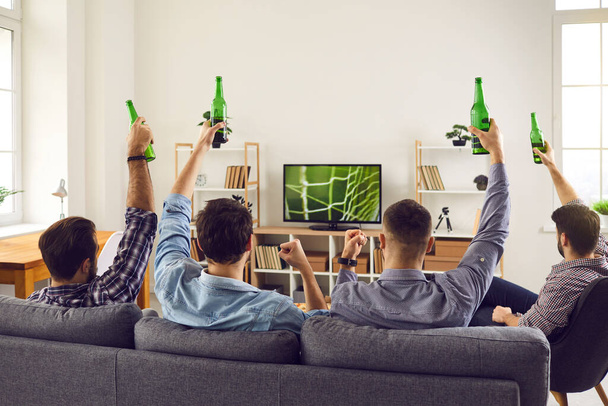 Um grupo de jovens amigos está assistindo um jogo de futebol em casa na TV, segurando uma garrafa - Foto, Imagem