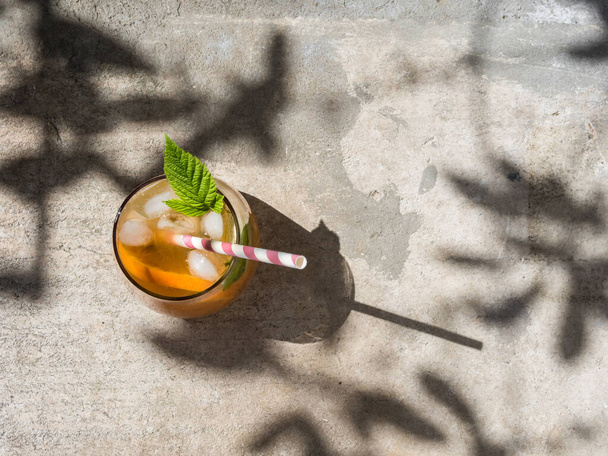 Cold orange cocktail with raspberry leaves, ice. Cocktail in glass with cocktail straw. View from above. The background is a natural concrete block with cement. Sunshine, shadow from tree foliage - Foto, imagen