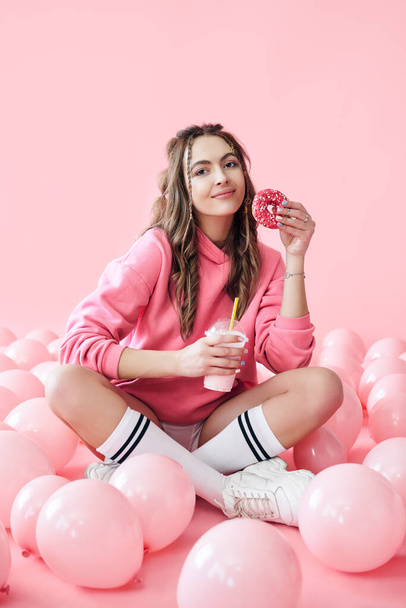 Smiling young woman with milkshake and donut in hands sitting on floor with many pink air balloons - Fotoğraf, Görsel