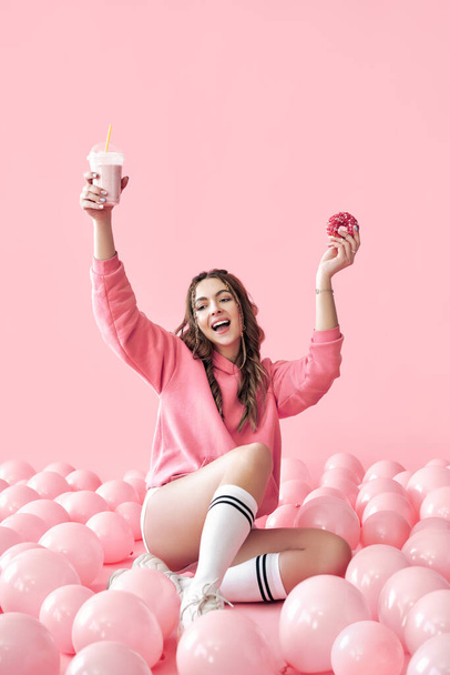 Happy young woman celebrate with hands up holding milkshake and donut - Fotó, kép
