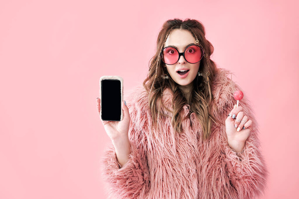 Surprised hipster woman holding blank screen mobile phone over pink background - Foto, imagen