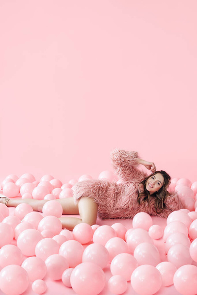 Fashion portrait of smiling pretty woman lying in many pink balloons on pink pastel background - Foto, imagen
