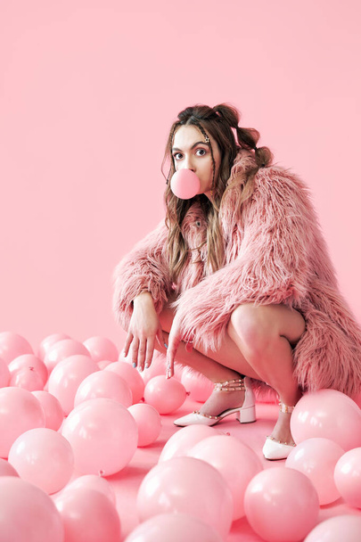 Funny hipster woman blowing bubble with chewing gum sitting in pink balloons background - Foto, Imagem