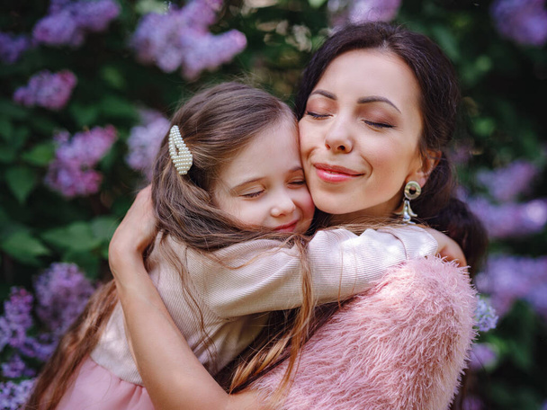 kobieta z córką W piękne różowe sukienki zabawy na świeżym powietrzu w wiosennym ogrodzie liliowym. Mama i dziecko uśmiechające się i przytulające. Rodzinne wakacje i wspólnota. Szczęśliwego Dnia Matki! - Zdjęcie, obraz