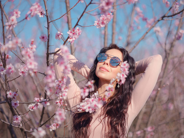 Çiçek açan kiraz ağacının yanında aynalı güneş gözlüklü güzel bir genç kadın. Yenilenme fikri ve konsepti, kişisel bakım, sağlık ve mutluluk - Fotoğraf, Görsel