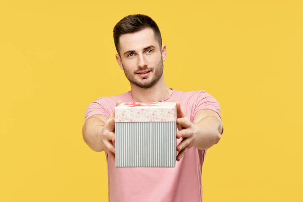 Handsome young man giving gift box looking to camera on yellow studio background - Photo, Image