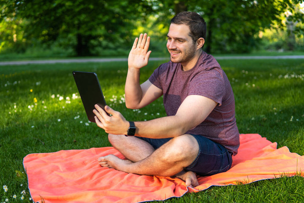 Foto de un hombre joven y atractivo sentado en una toalla en el parque haciendo una videollamada a través de una tableta con un amigo y disfrutando de un día de verano - Foto, imagen