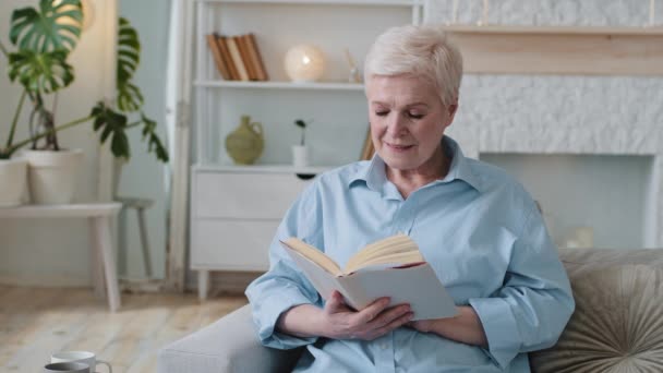 Entspannt liest eine alte Frau einen Bestseller aus dem Buch, sitzt auf dem Sofa und verbringt das gemütliche Wochenende zu Hause. Glückliche Seniorin genießt Freizeit mit Hobby und ruht sich auf Couch im Wohnzimmer aus. - Filmmaterial, Video