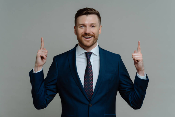 Happy businessman in suit pointing up with fingers and smiling at camera - Photo, Image