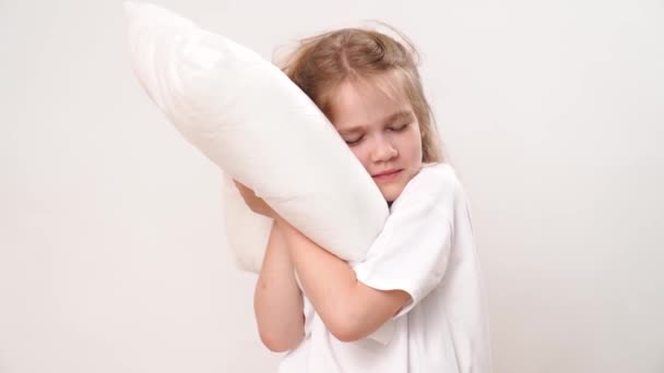 可愛いシャギー眠い子供の女の子は白い背景に枕を抱いています。幼児期に学校や幼稚園に通っていました。快適な寝具. - 映像、動画