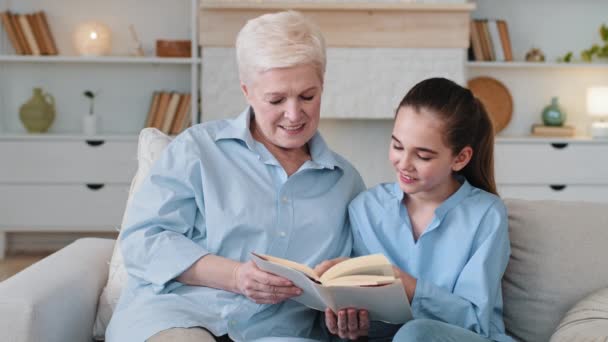Smart girl granddaughter reading with grandma at home. Senior grandmother babysitter or elderly private tutor help to schoolgirl with homework explains material sits together on couch hobby concept - Footage, Video