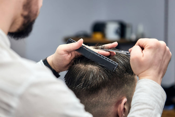 Professional barber making haircut to young man using scissors and comb at barbershop. Close-up, selective focus - Foto, imagen