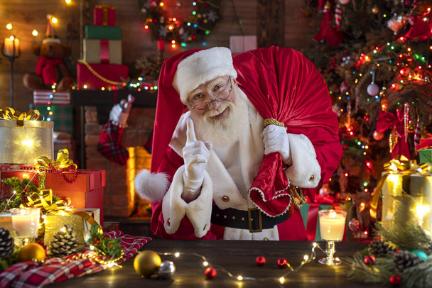 Santa Claus with a bag with presents gifts near fireplace and Christmas tree. Festive interior inside wooden house, New Year's cheerful mood Spirit of Christmas. Senior man with real white beard cosplay Father Christmas. - Photo, Image