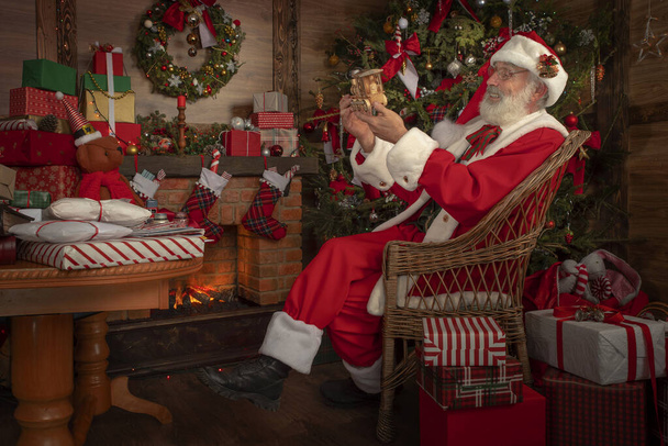 Weihnachtsmann bastelt Spielzeug in der Werkstatt im festlichen Interieur des Holzhauses, Neujahrsstimmung. Senior Mann mit echtem weißen Bart cosplay Weihnachtsmann. - Foto, Bild