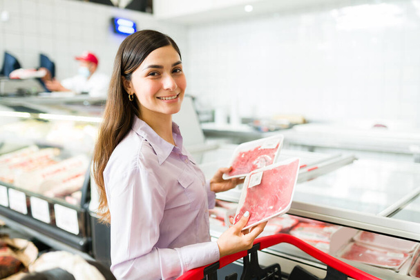 Schnelle Einkaufstour im Supermarkt. Schöne lateinische Frau hält verpacktes rohes Fleisch in der Hand und wählt frische Lebensmittel, die sie im Lebensmittelgeschäft kaufen kann - Foto, Bild