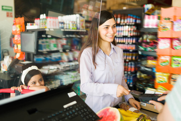 Female customer smiling and paying with a credit card at the supermarket. Good-looking woman and kid at the checkout counter buying food  - Photo, Image