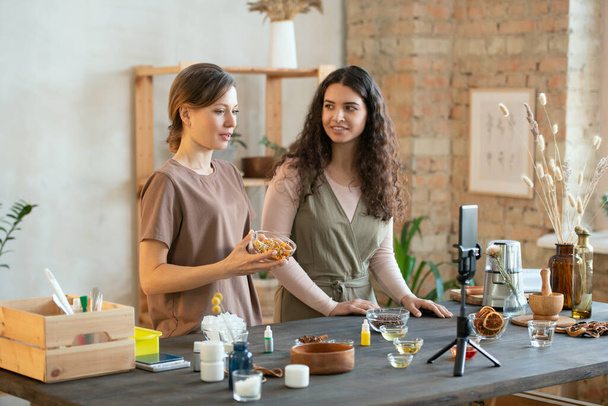 İki genç kadının elleri, kozmetik ürünler üretmek için malzemelerle birlikte masada dururken tahtalarda sert sabun kütlelerini kesiyor. - Fotoğraf, Görsel