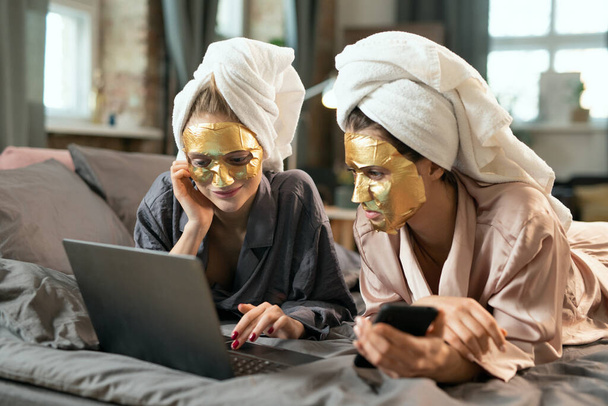 Dos hembras jóvenes en pijama de seda, toallas en la cabeza y máscaras cosméticas doradas en rostros relajantes en la cama y desplazándose en smartphone - Foto, Imagen
