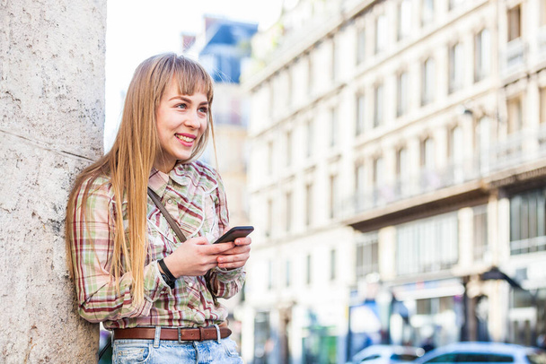 Happy young woman using her phone in Paris - Portrait of a smiling blonde girl standing on the side of the street and looking away - Travel and lifestyle concepts - Photo, Image