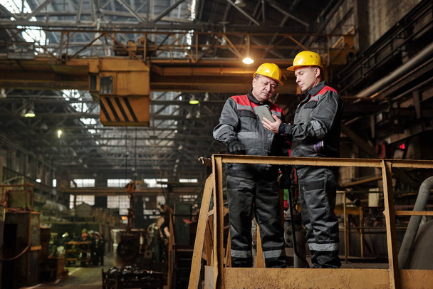 Two manual workers in workwear using digital tablet and discussing work while standing in the plant - Photo, Image