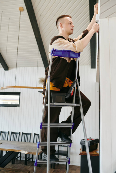 Νεαρός μάστορας με στολή σκαρφαλώνει σε σκάλα ενώ κάνει τη δουλειά του - Φωτογραφία, εικόνα
