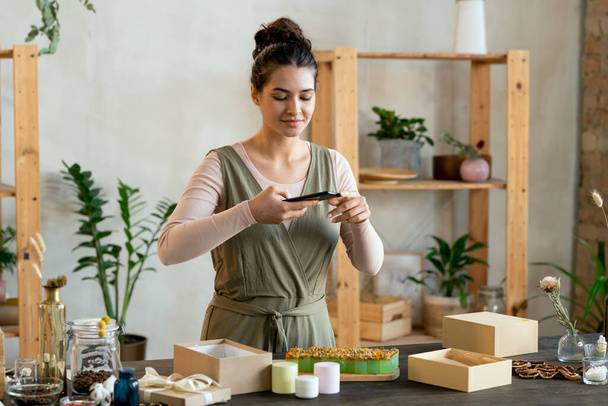 Young woman with smartphone shooting video while standing by table with fresh aromatic handmade soap and candles before packing them - Foto, afbeelding