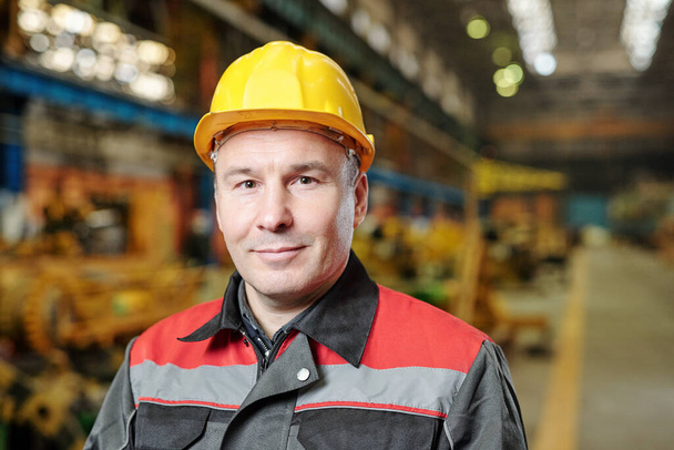 Portret van volwassen handarbeider in helm en werkkleding op zoek naar camera tijdens het gebruik van tablet pc in de fabriek - Foto, afbeelding