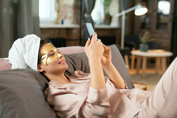 Joven hermosa mujer en pijama de seda, toalla en la cabeza y máscara cosmética dorada en la cara relajante en la cama y haciendo selfie - Foto, Imagen