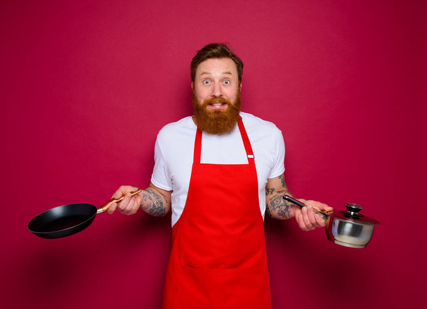 испуганный шеф-повар с бородой и красный фартук готовит с кастрюлей и кастрюлей - Фото, изображение