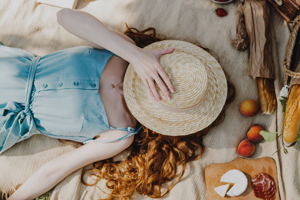 Young woman in blue dress hiding face from sunlight under straw hat while lying on the grass in park. - 写真・画像