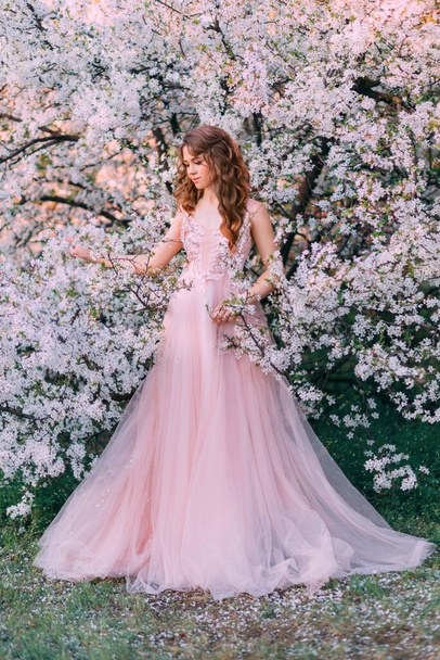 Young red-haired girl princess in pink evening tulle fluffy dress. Elegant romantic woman walking in blooming cherry garden. Enjoyment of Spring Nature. Green trees white flowers. Fantasy vintage lady - 写真・画像