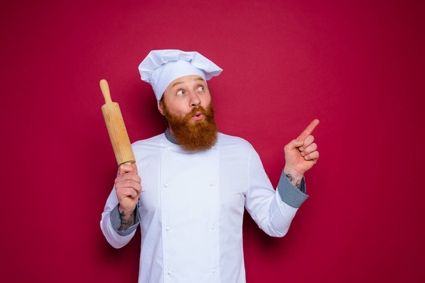 užaslý kuchař s plnovousem a červenou zástěrou šéfkuchař drží dřevěný váleček - Fotografie, Obrázek