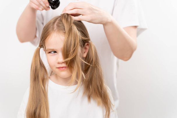 woman combs and does the hair of a funny little girl on a white background. Mommy is a hairdresser. saving money at a beauty salon. shampoos and cosmetics for children's hair. - Photo, Image