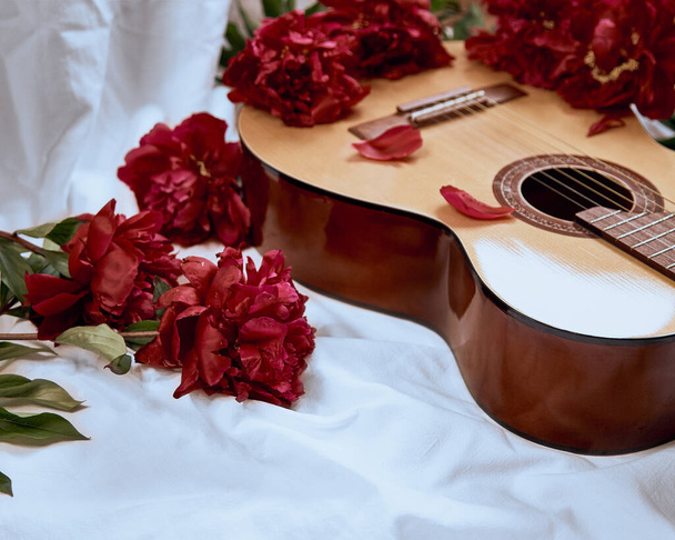 アコースティック木製のギターは、赤い牡丹に囲まれた白いシートにあります。音楽アルバム、音楽コンセプト、ギター演奏、ロマンチックなセレナーデのカバー - 写真・画像