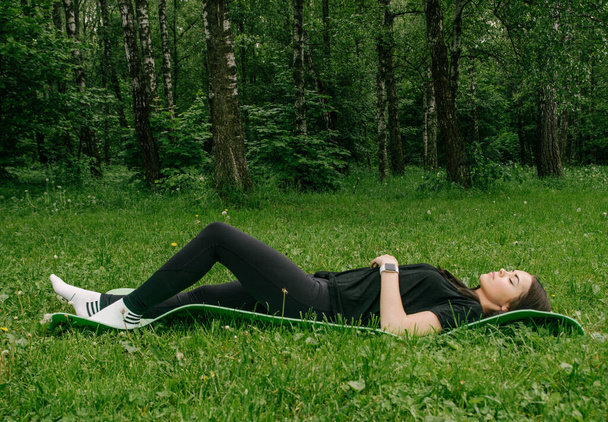 a girl in a black T-shirt and leggings lies on a sports mat and rests after classes in the green - Фото, зображення