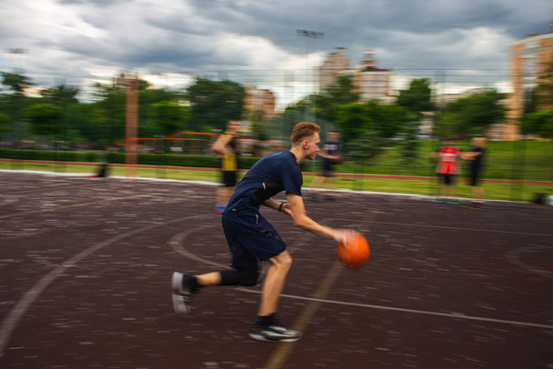 Genç kızıl saçlı bir adam yüksek hızda koşuyor ve top sürüyor. Gün içinde sokaktaki bir spor sahasında hareket bulanıklaştı. - Fotoğraf, Görsel