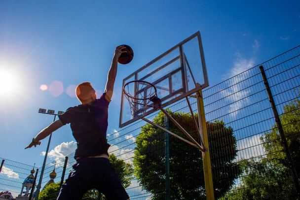 Joven pelirrojo con una camiseta azul oscuro lanza una pelota en movimiento en un aro de baloncesto contra un cielo azul al aire libre - Foto, Imagen