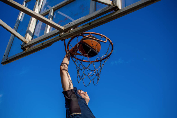 Jeune homme roux en t-shirt bleu foncé lance une balle en mouvement dans un panier de basket contre un ciel bleu en plein air - Photo, image