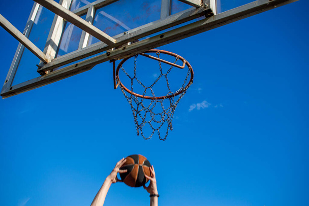 Νεαρός κοκκινομάλλης με σκούρο μπλε μπλουζάκι ρίχνει μια μπάλα σε κίνηση σε ένα στεφάνι μπάσκετ σε έναν εξωτερικό μπλε ουρανό - Φωτογραφία, εικόνα