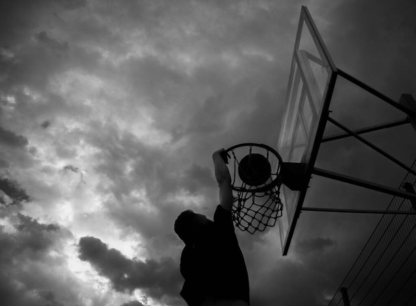 Σιλουέτα ενός ανθρώπου που ρίχνει μια μπάλα σε ένα στεφάνι μπάσκετ στο δρόμο ενάντια σε έναν ουρανό με σύννεφα σε μαύρο και άσπρο χρώμα - Φωτογραφία, εικόνα