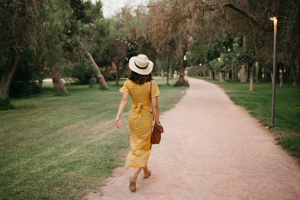 Фото брюнетки в жёлтом платье и шляпы, идущей по песчаной дорожке в Валенсийском парке вечером. - Фото, изображение